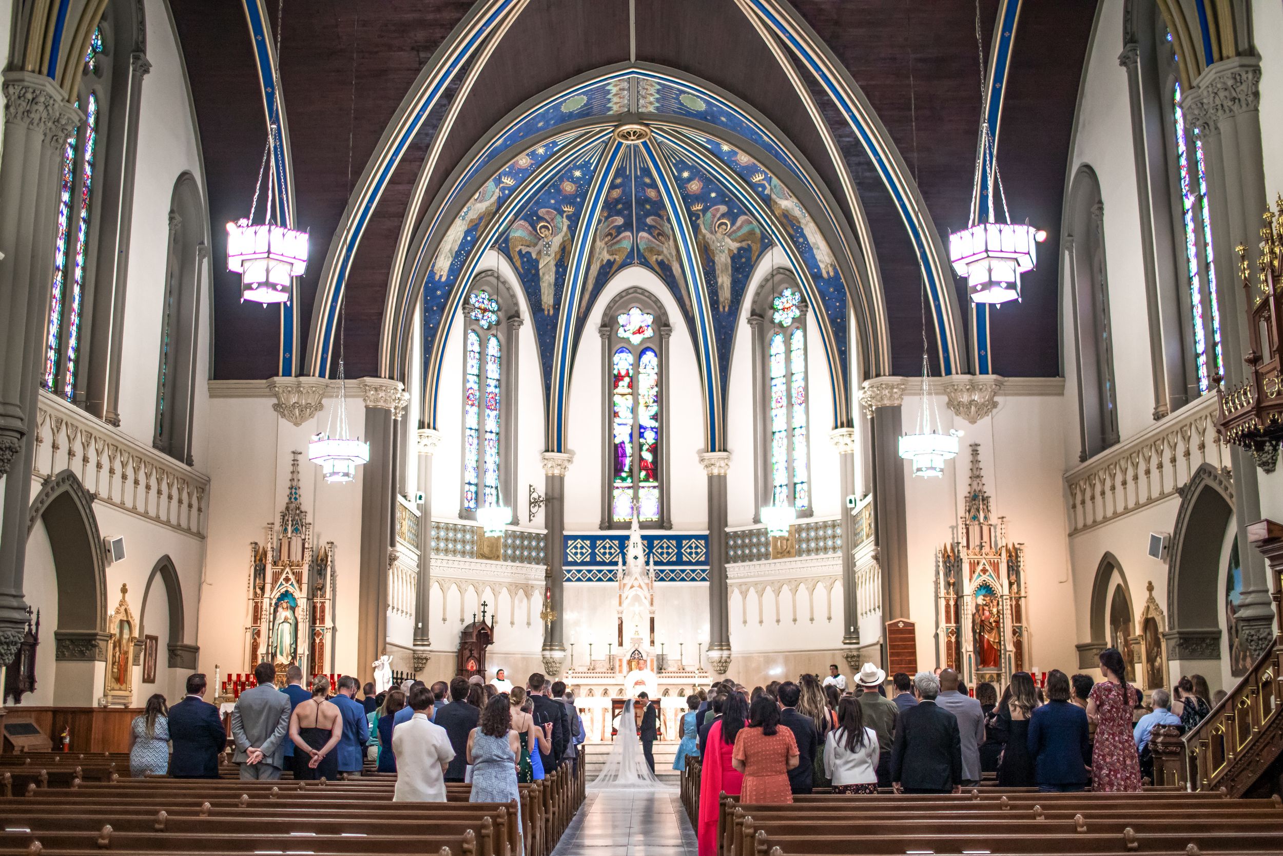 Interior view of a Catholic church with vaulted blue ceiling, stained glass windows and congregation gathered for service.