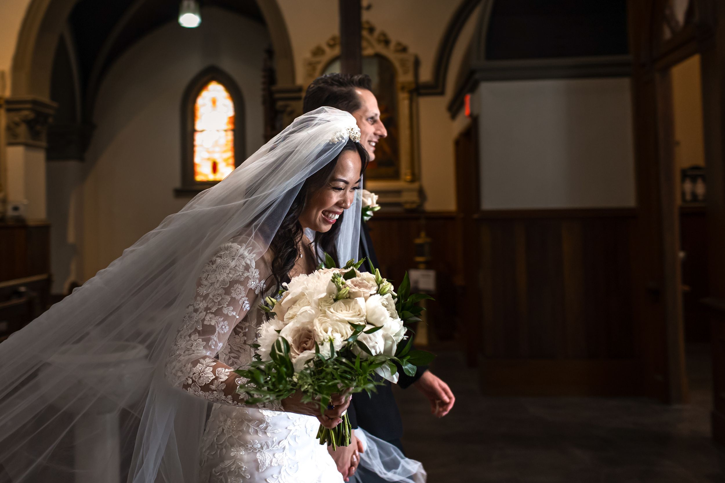 A bride and groom share a tender moment in a dimly lit church with gothic arched windows and warm lighting.