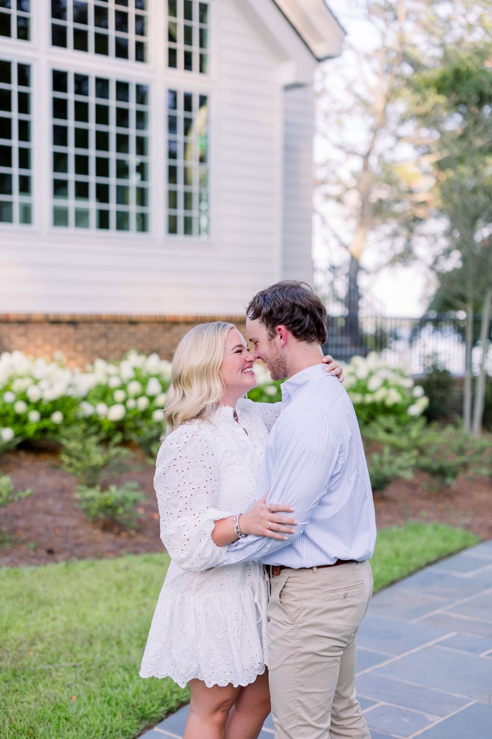 A romantic couple shares a tender embrace on a stone patio next to white hydrangea bushes and a brick wall.