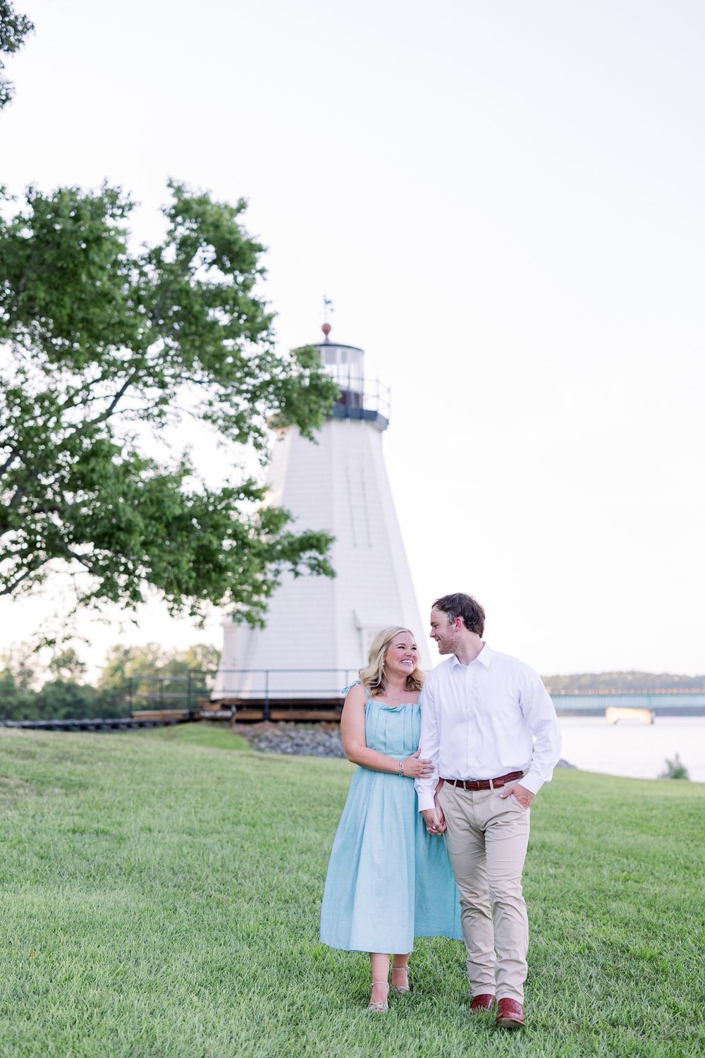 A couple in light blue and khaki outfits walk together on a grassy hillside near a lighthouse during golden hour.
