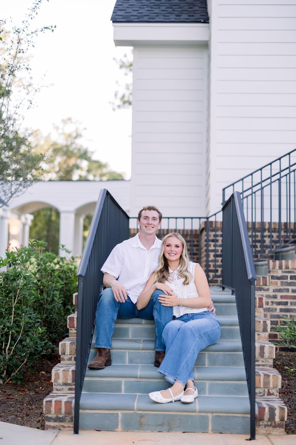 Couple sitting and laughing together on outdoor gray concrete steps with white house and greenery in background.