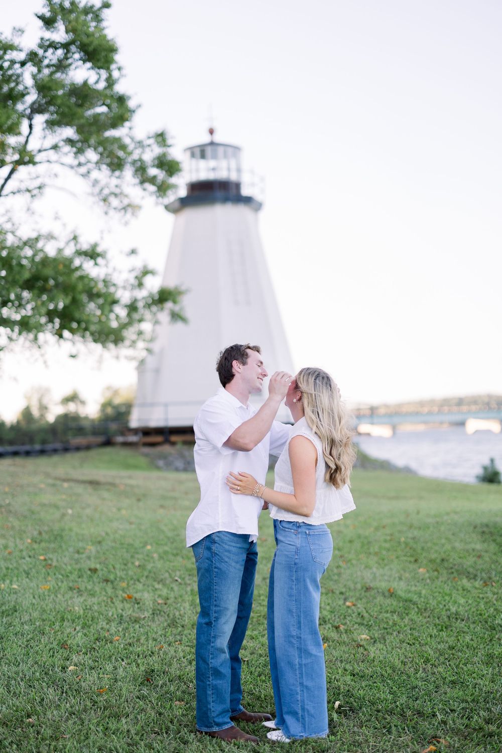 Couple sharing romantic moments on grassy hill with white lighthouse visible in background.