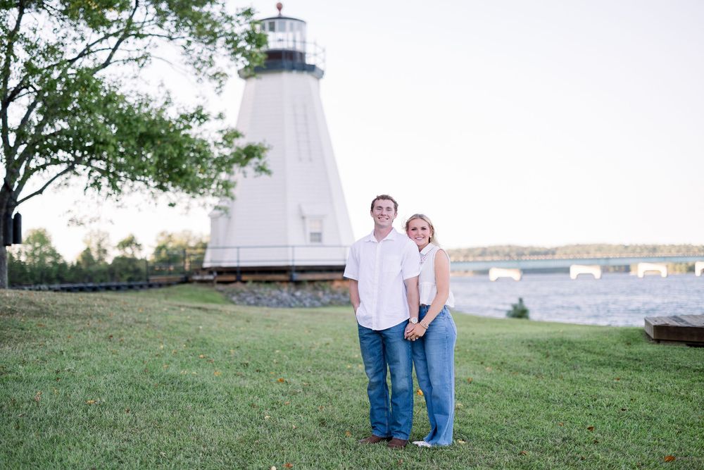 A couple poses in front of a white lighthouse during an outdoor photo session at sunset.