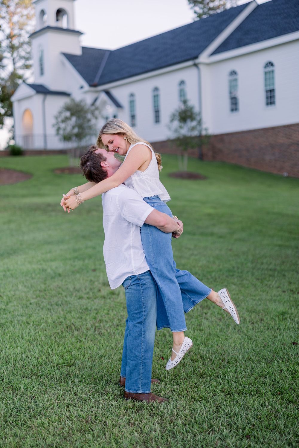 A couple dances and spins together on a grassy lawn with a white church building visible in the background.