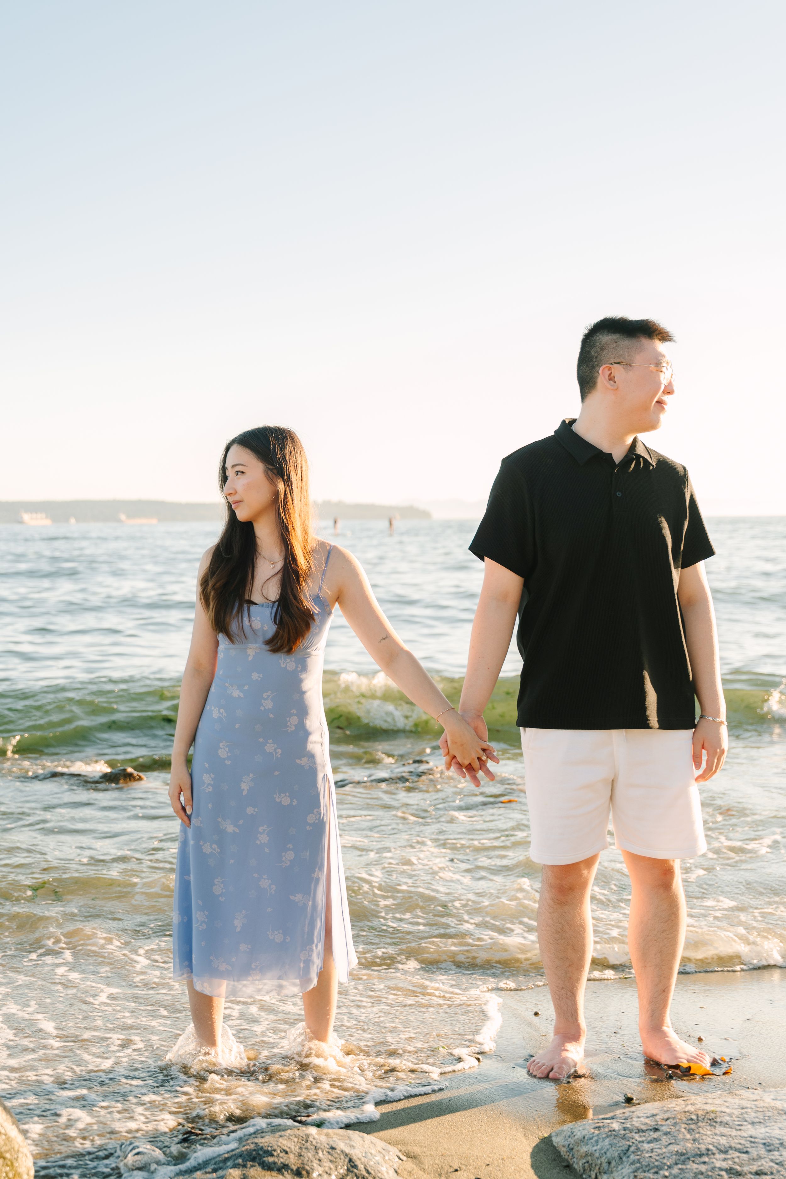 A couple walks hand in hand along a sunny beach shoreline with waves gently lapping at their feet.