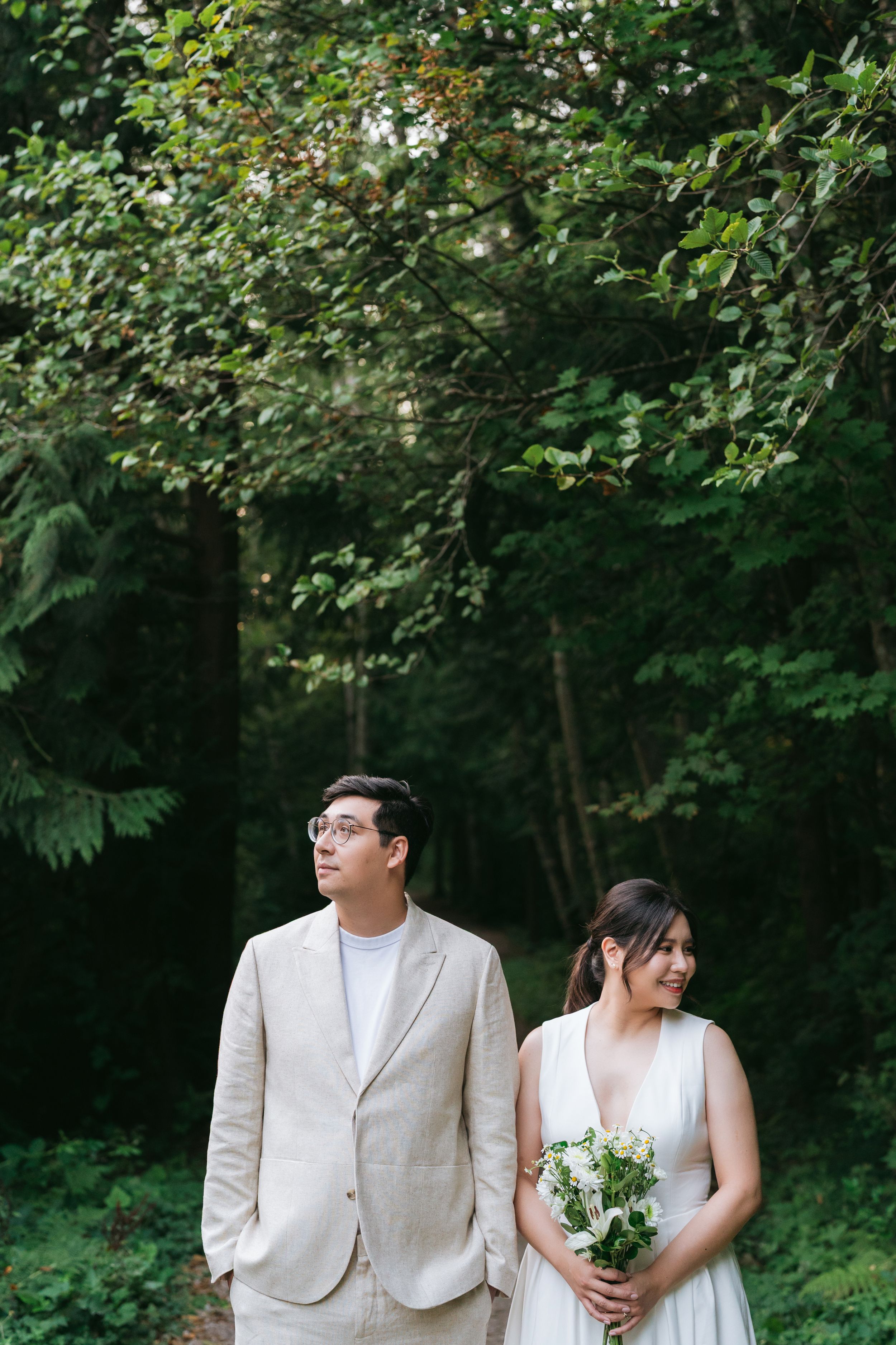 A couple in wedding attire stands in a lush green forest setting, with the bride holding a simple bouquet.