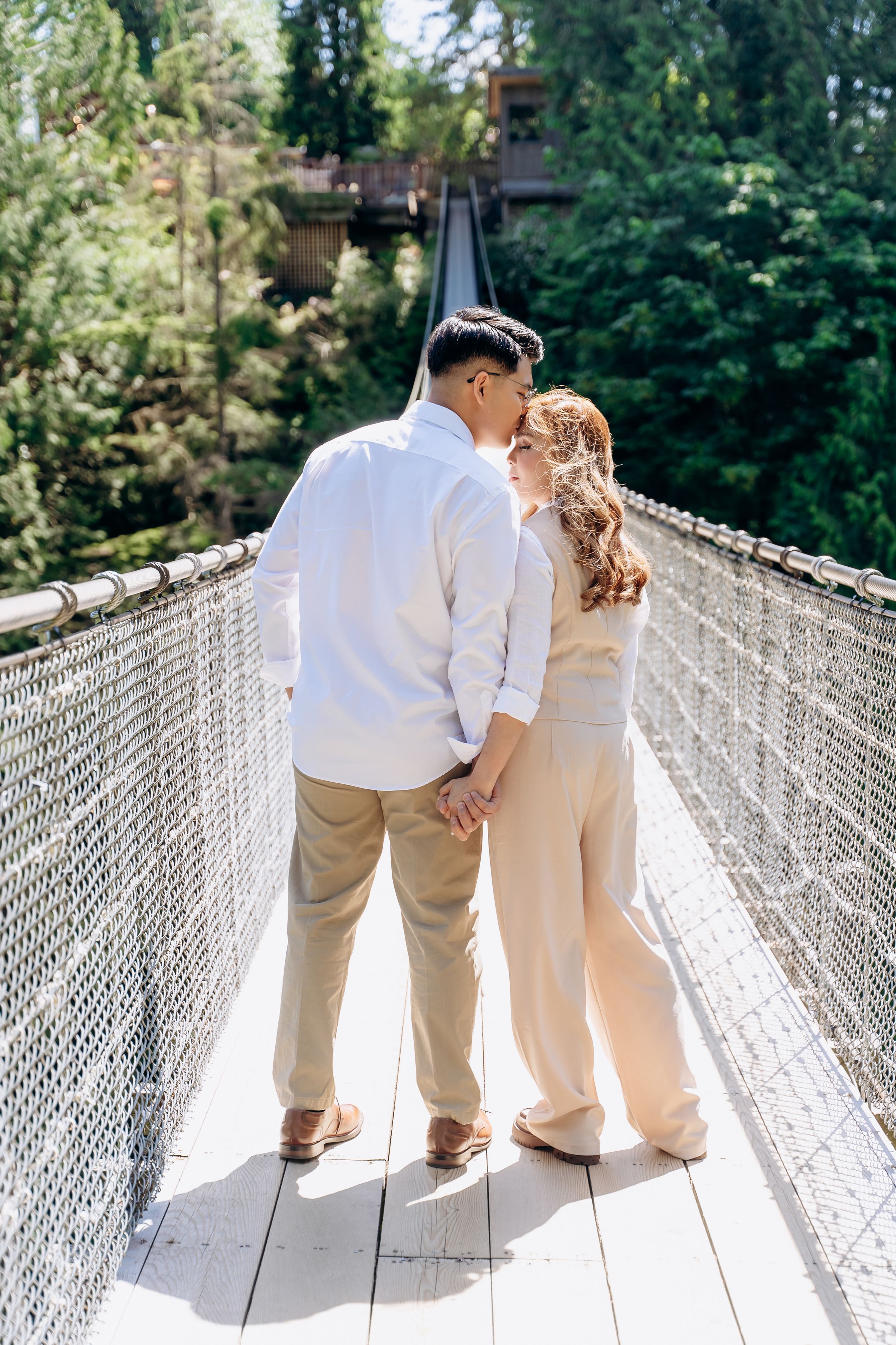 A couple shares an intimate moment on a scenic suspension bridge surrounded by evergreen trees in sunlit forest.