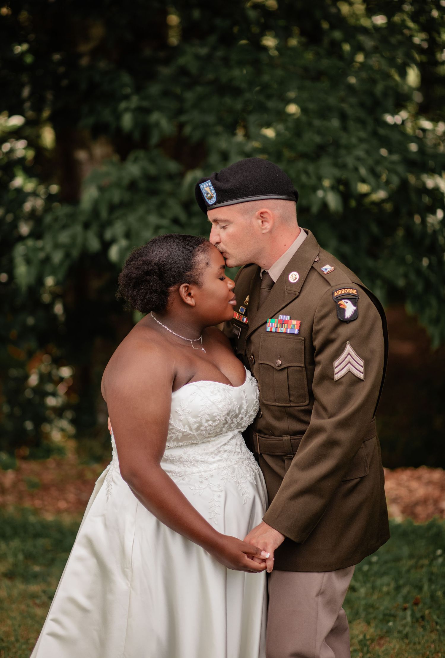 A couple shares a romantic moment outdoors during a military wedding celebration, with the bride in white and groom in uniform.