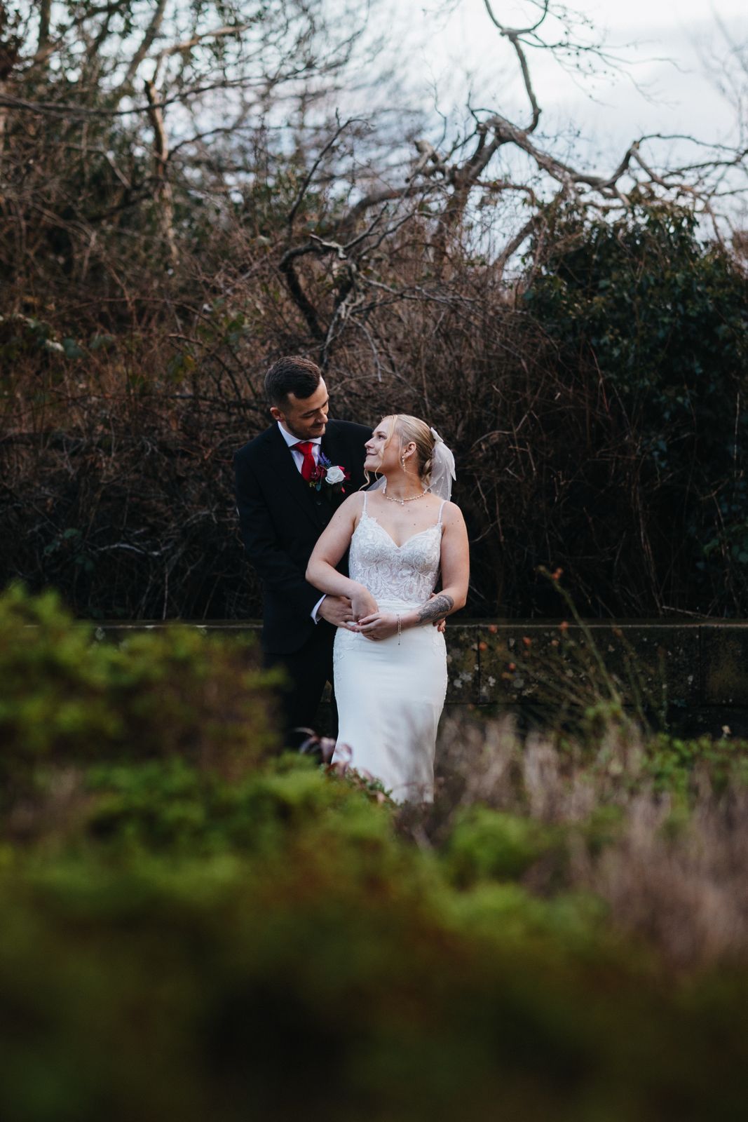 A couple in wedding attire walks together through a scenic outdoor setting with bare winter trees in the background.
