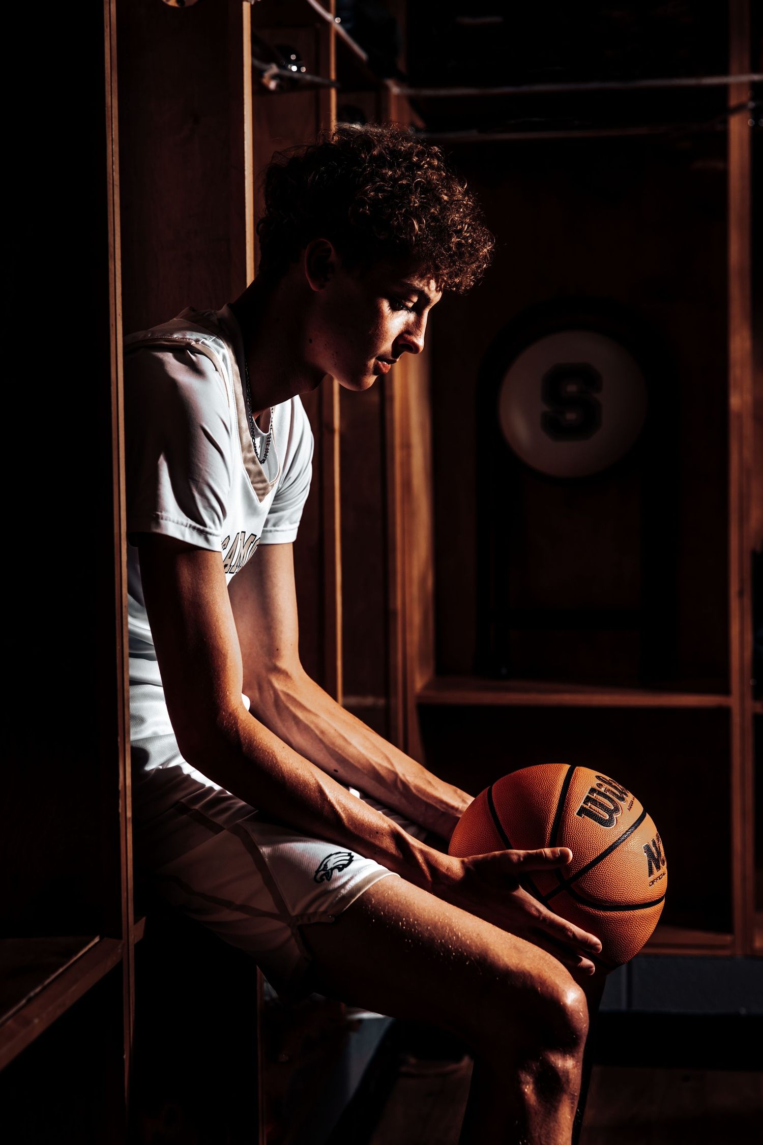 A basketball player sits in dramatic lighting holding a ball while wearing white and brown athletic attire.