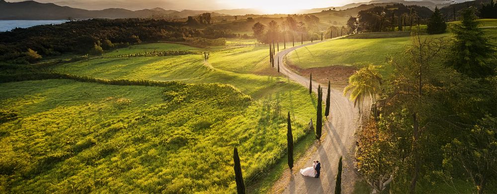 Aerial view of a bride and groom walking along a scenic countryside road at golden sunset with rolling green hills.