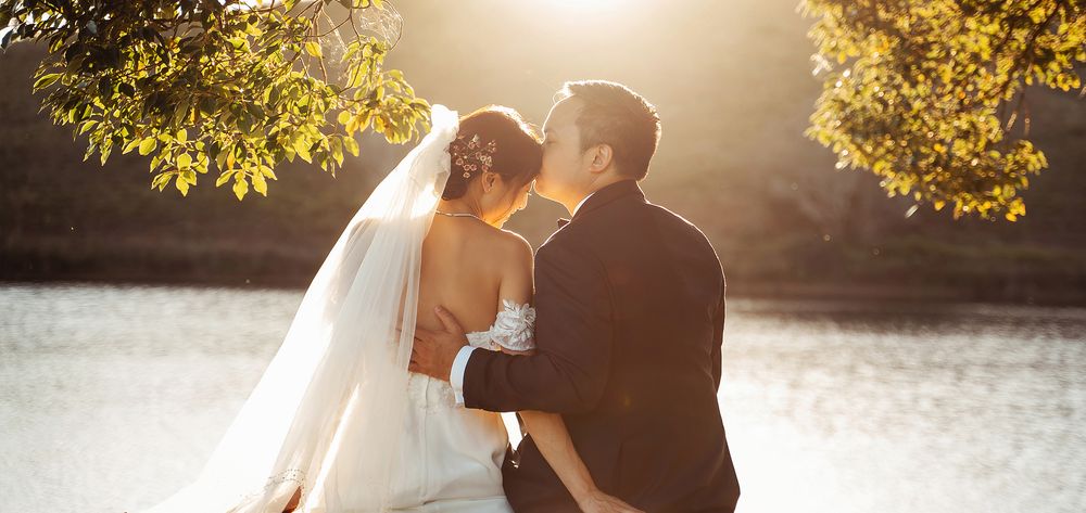 Bride and groom share a tender kiss by a sunlit lake under golden autumn foliage.