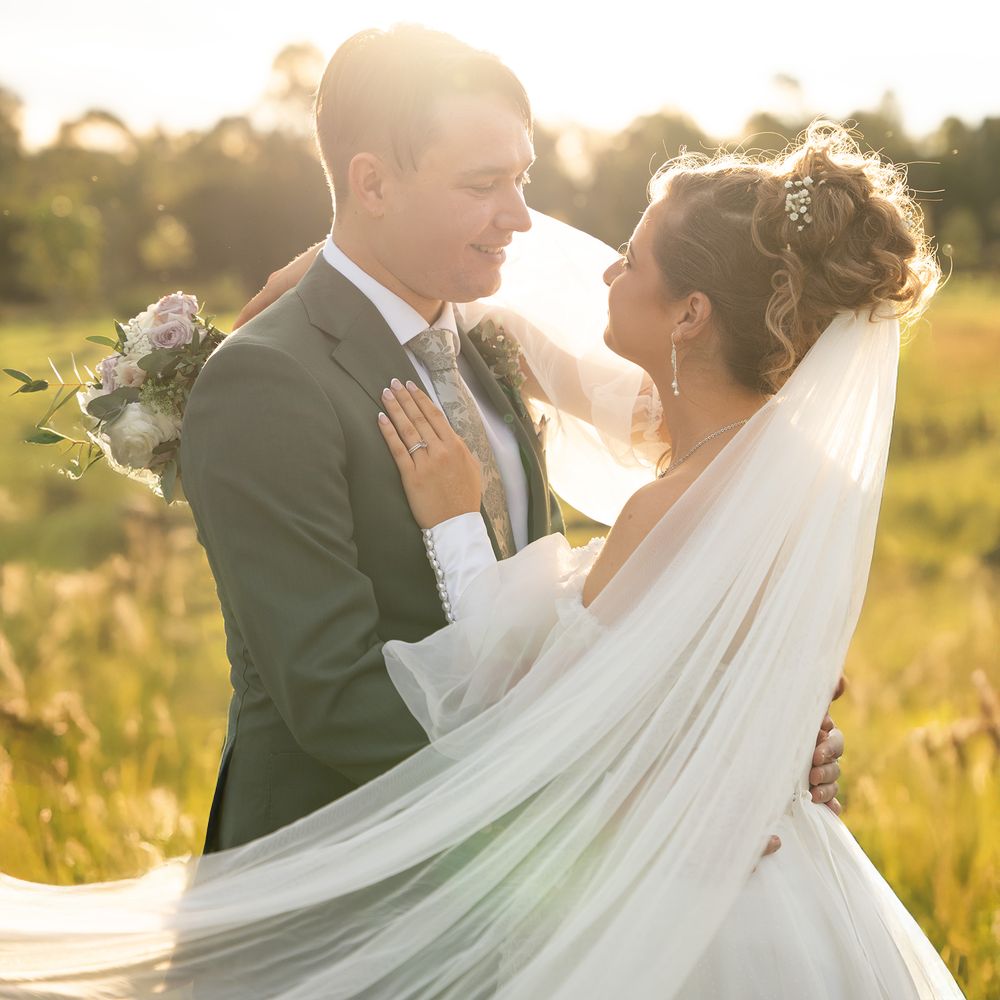 Bride and groom share a romantic moment in a golden sunlit field, veil flowing in the breeze.