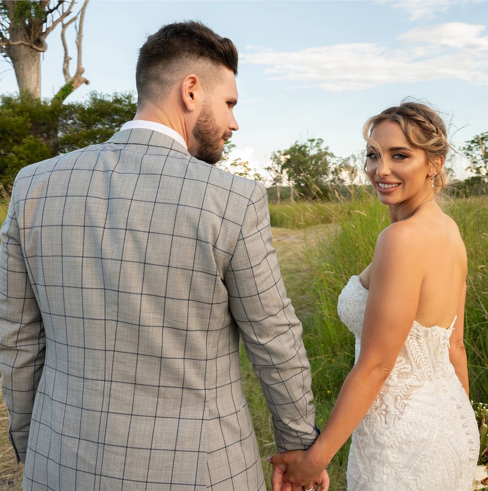 Bride smiling over shoulder while holding groom's hand on scenic outdoor wedding day path.