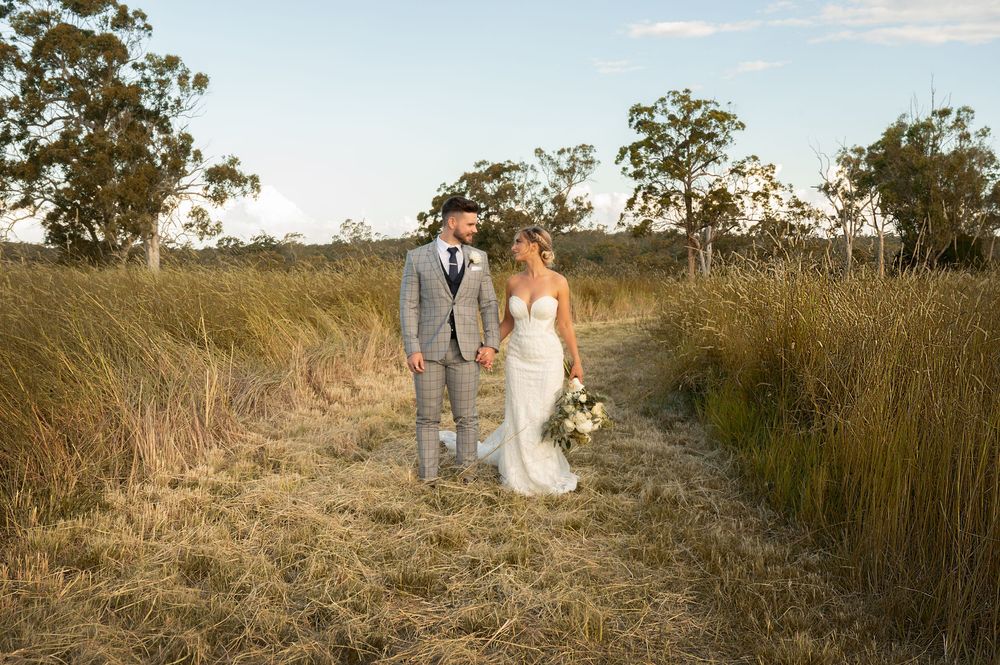 Bride and groom standing together in golden dry grass field surrounded by Australian bush trees.