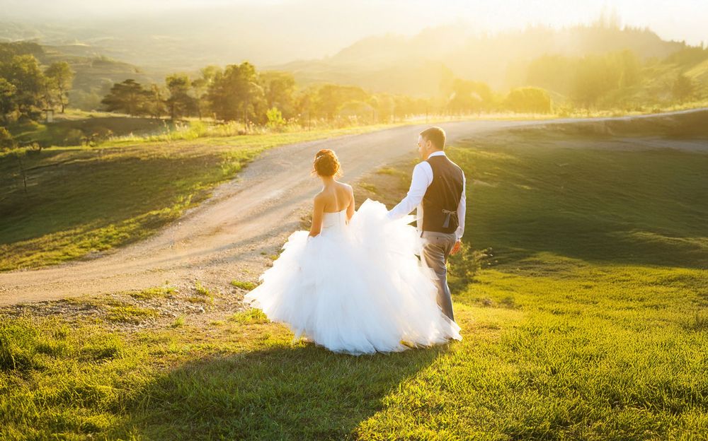 A bride and groom walk hand in hand through a sunlit green countryside on their wedding day.