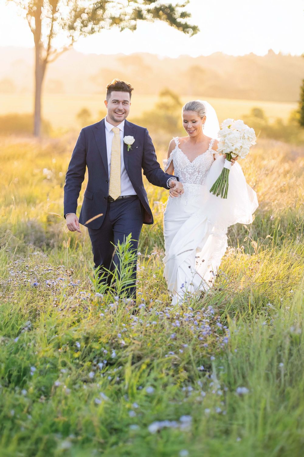 Bride and groom walking hand in hand through wildflower field bathed in golden sunset light.