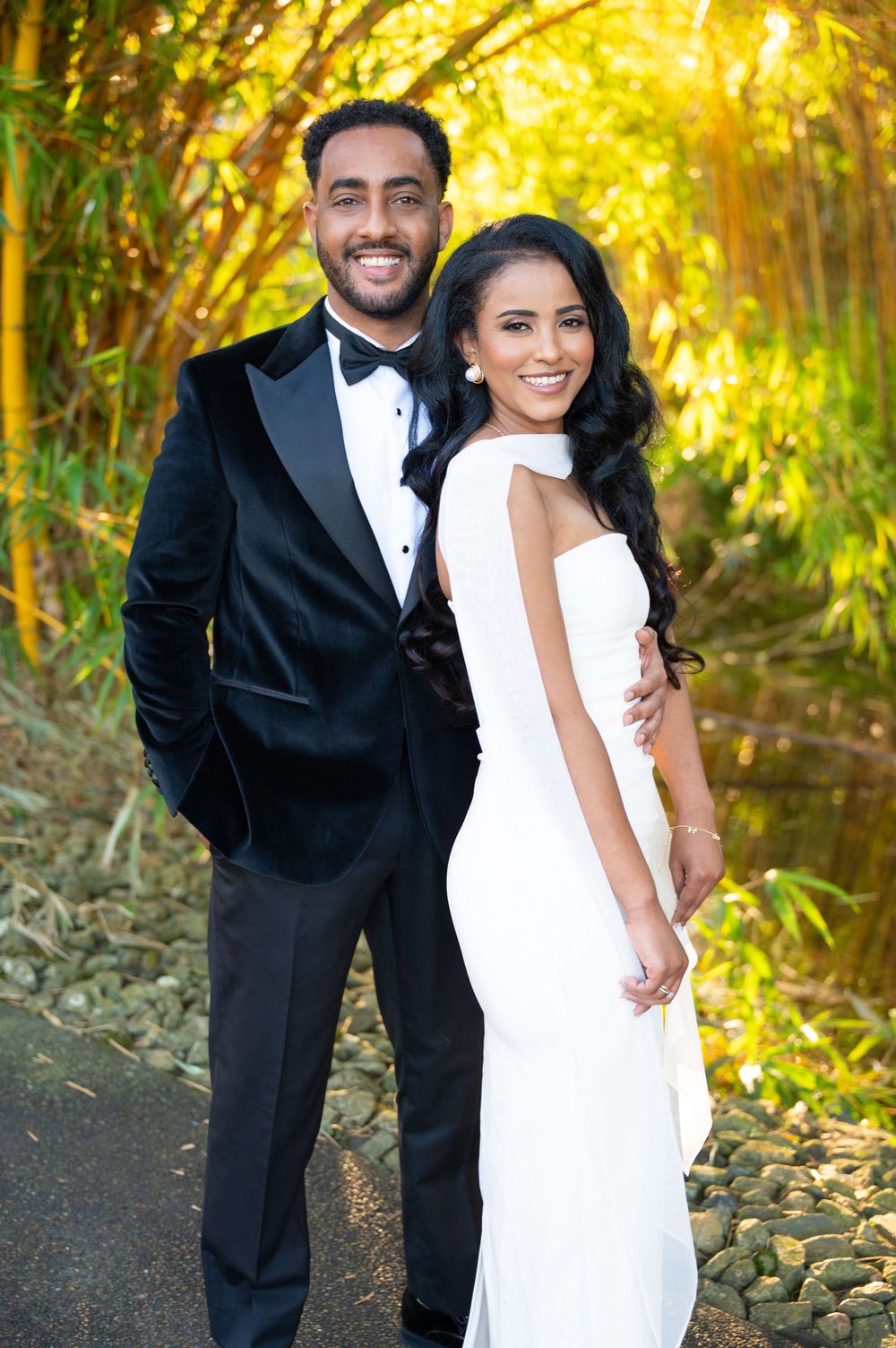 Elegant couple posing in formal wedding attire, man in black tuxedo and woman in white gown, surrounded by lush greenery.