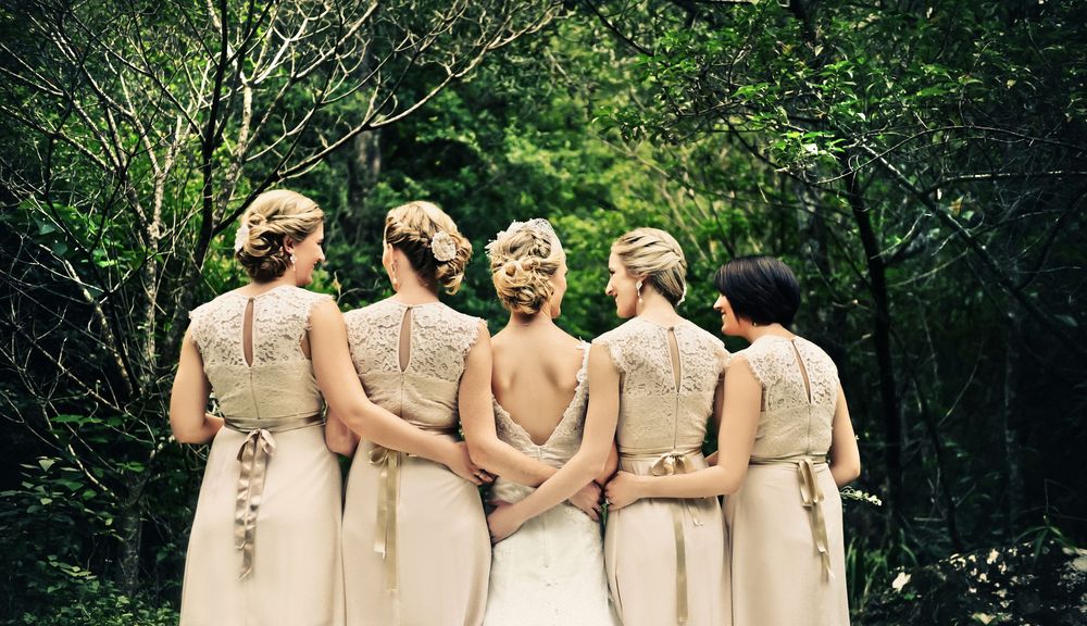 Four bridesmaids in champagne gowns standing together with backs to camera in lush green forest.