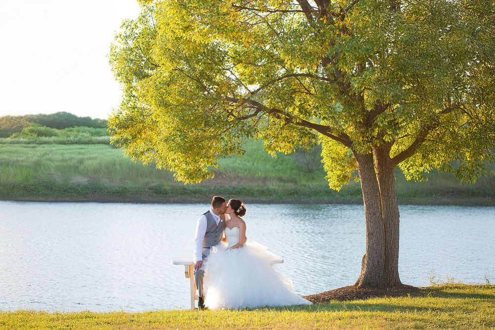 A bride and groom share a romantic kiss under a large green tree by a serene lake on their wedding day.