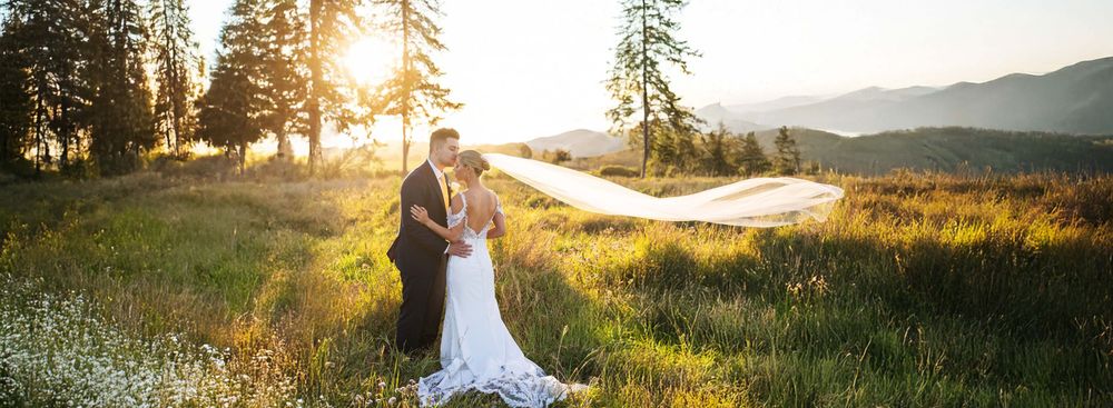 Bride and groom embrace in golden sunset light in a scenic mountain meadow with veil flowing.