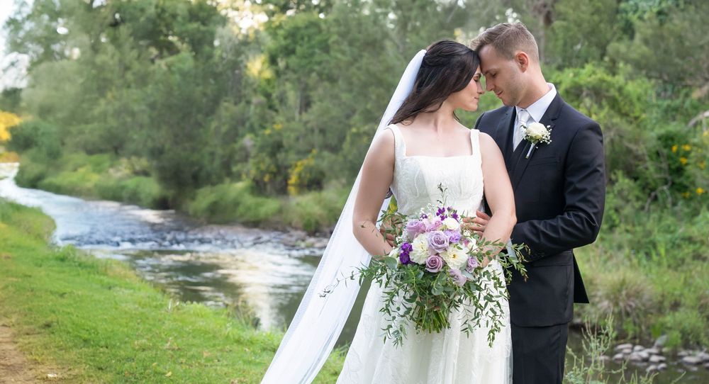 Bride and groom sharing a romantic moment beside a flowing river with lush green trees.