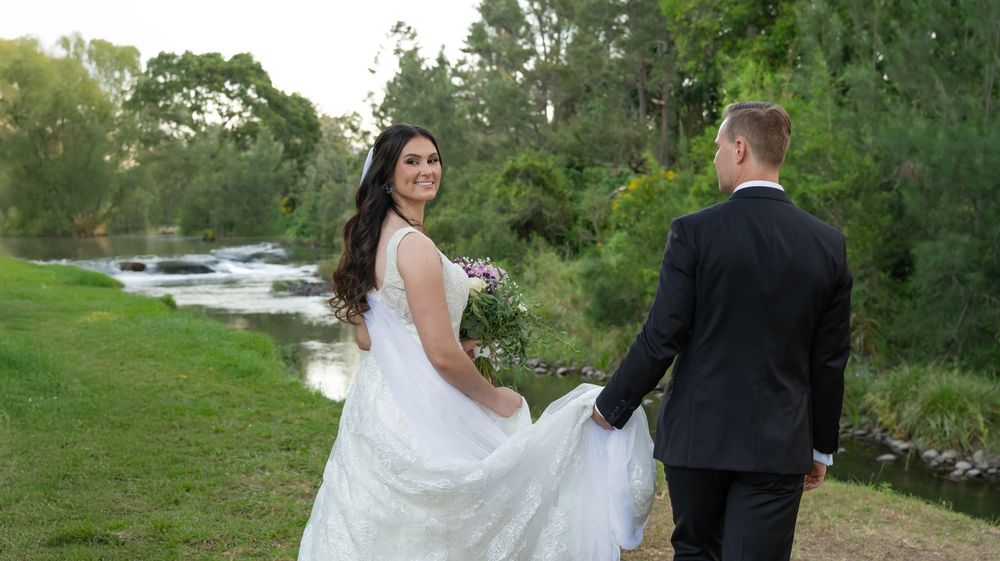 Bride and groom holding hands walking by a scenic river surrounded by lush greenery.