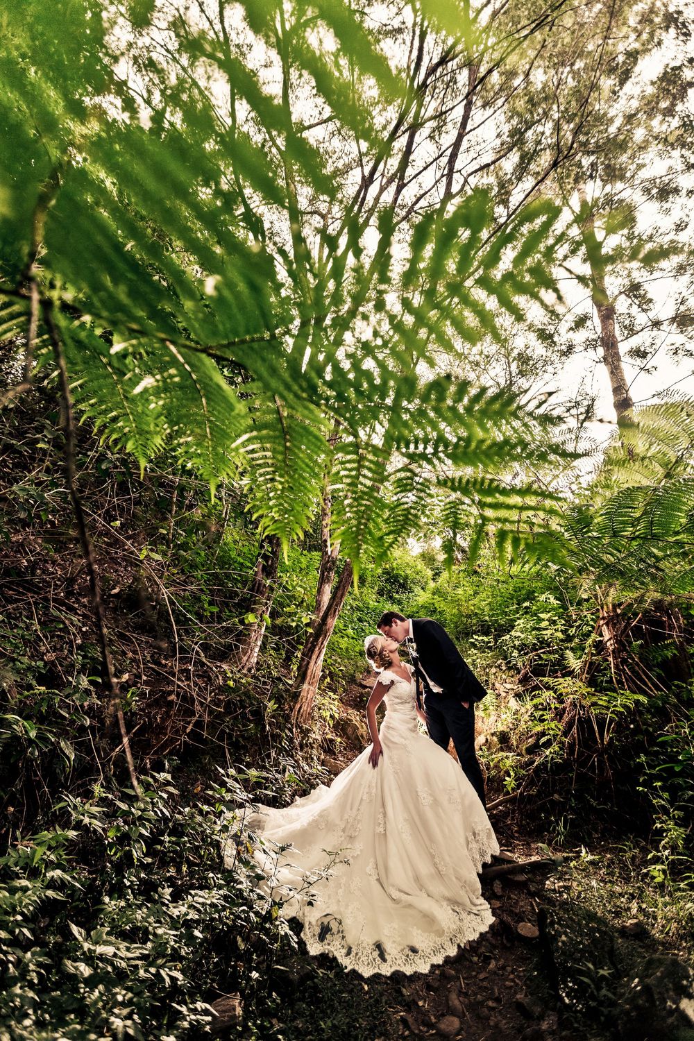 Bride and groom sharing romantic kiss beneath lush tropical ferns in enchanting forest setting.