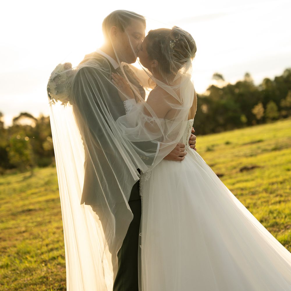 Bride and groom share a romantic kiss bathed in golden sunset light on a lush green farm.