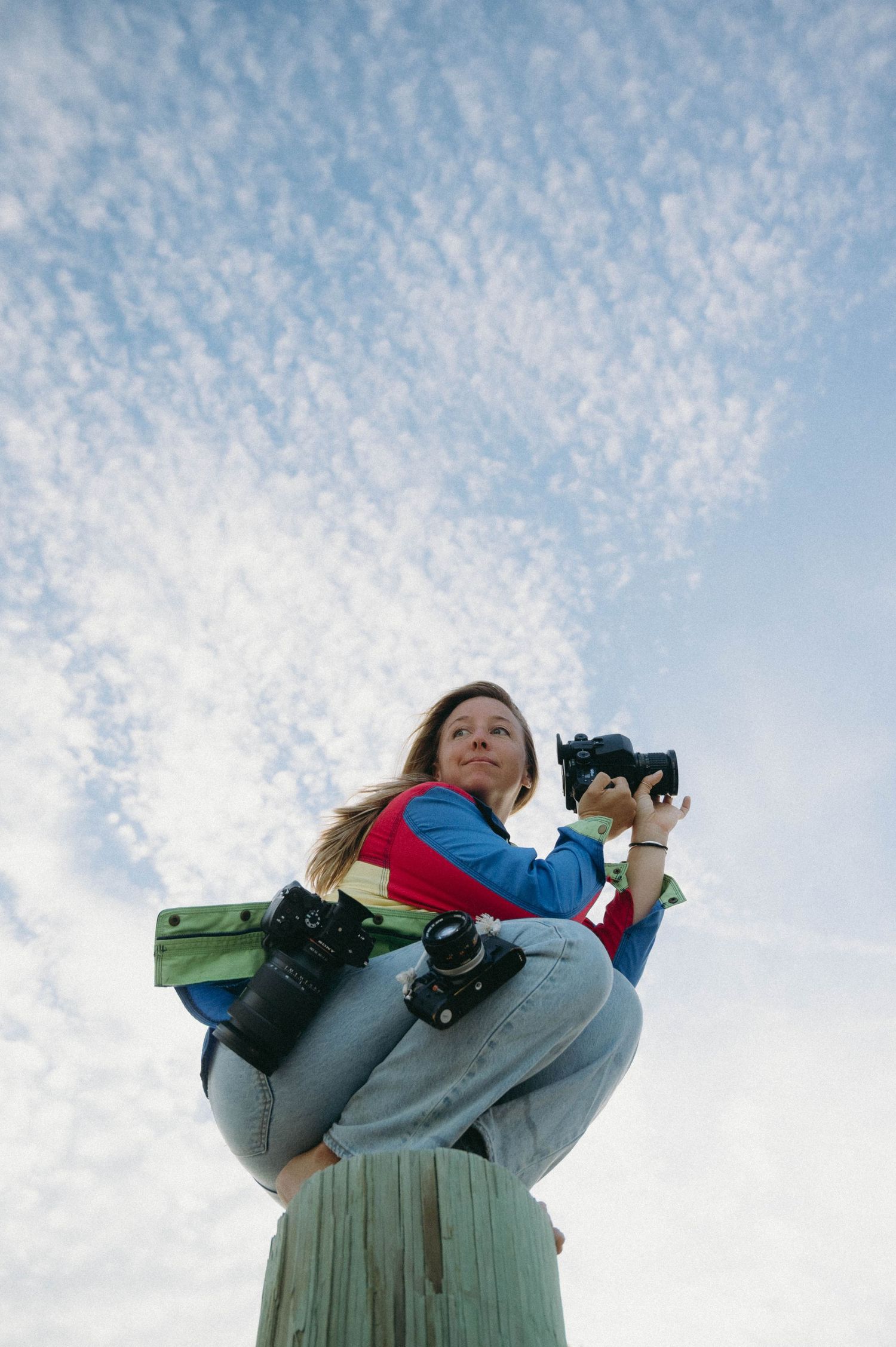 Person sitting atop a wooden post against a cloudy blue sky while holding a camera.