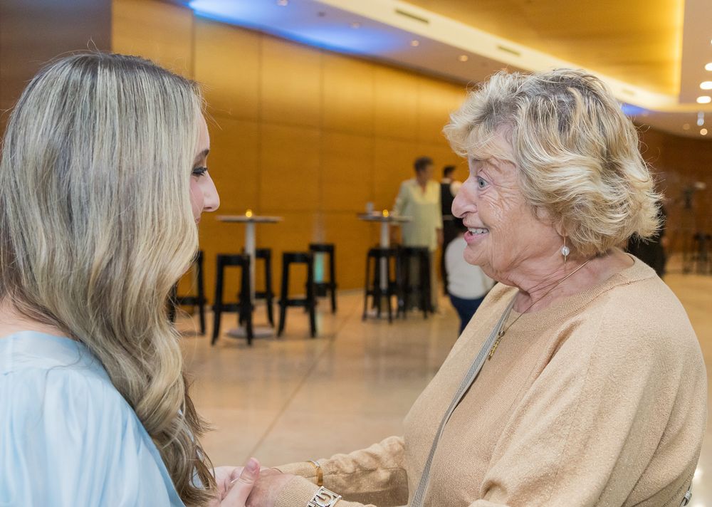 Two people having an animated conversation in a lobby area with warm lighting and bar stools visible in the background.