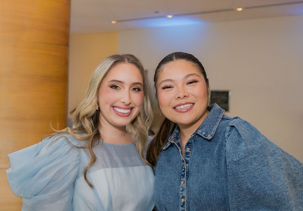 Two friends smile together indoors with one wearing a light blue top and another in denim.
