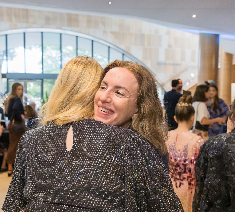 Two people share a joyful embrace at an indoor social gathering with other attendees visible in the background.