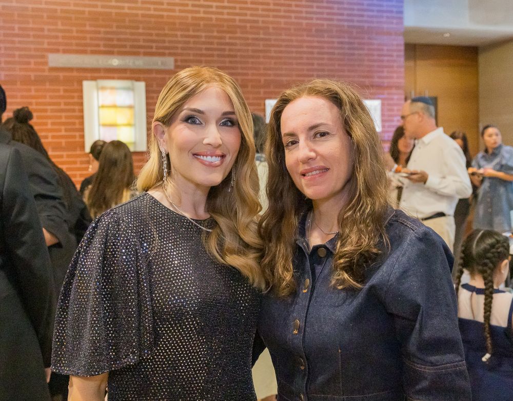 Two women pose together at an indoor event, one wearing a sparkly dark top and one in denim.