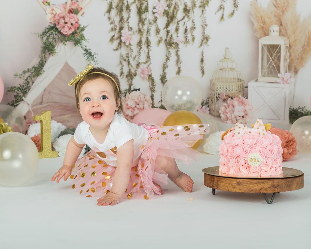 A happy baby girl in pink tutu sits near birthday cake during first birthday photoshoot.