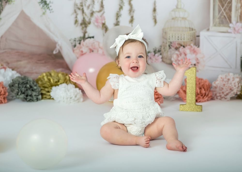 A joyful baby girl in white lace romper claps hands at her first birthday cake smash.