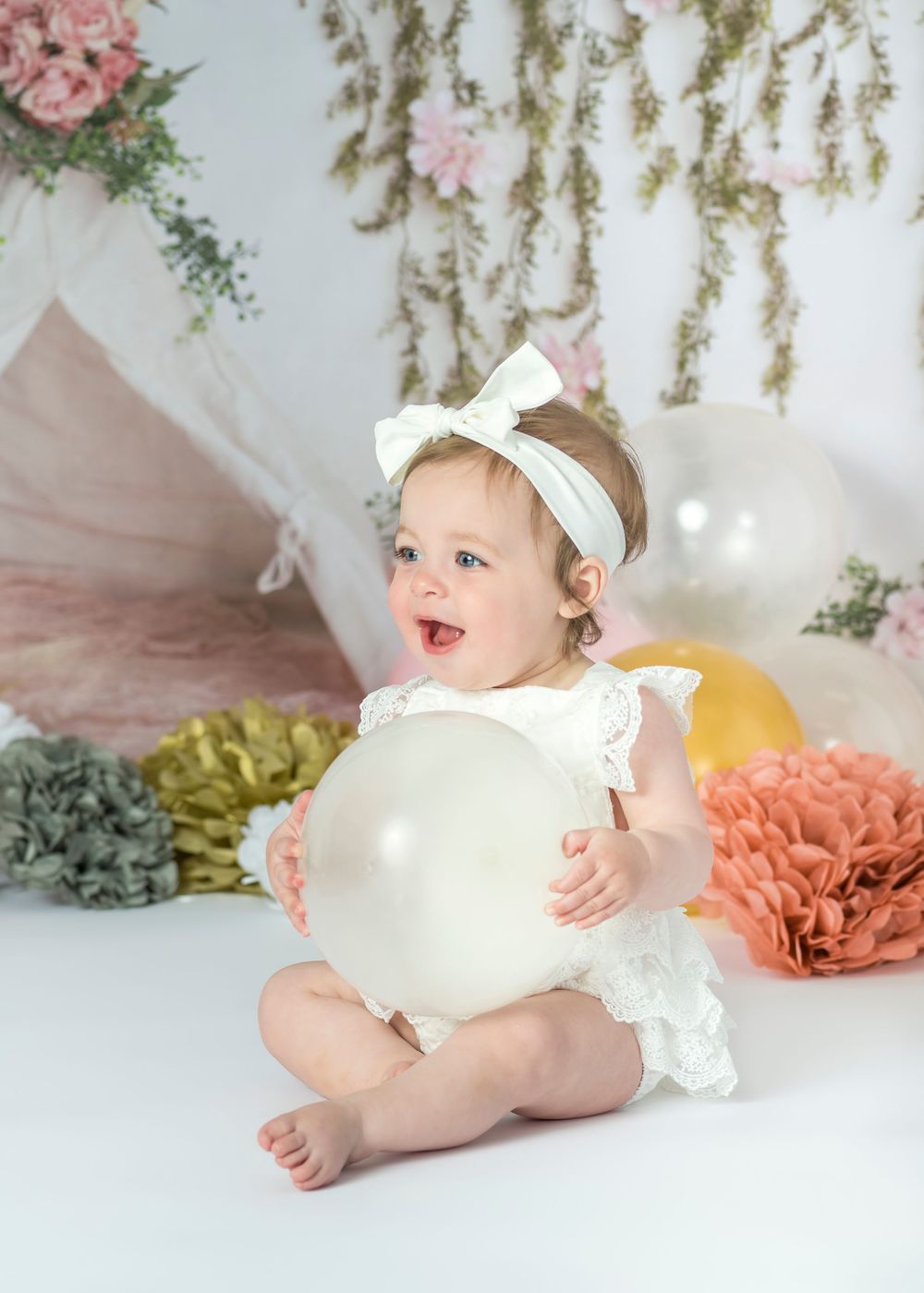 A happy baby girl in white lace romper holds a balloon at her first birthday photoshoot.