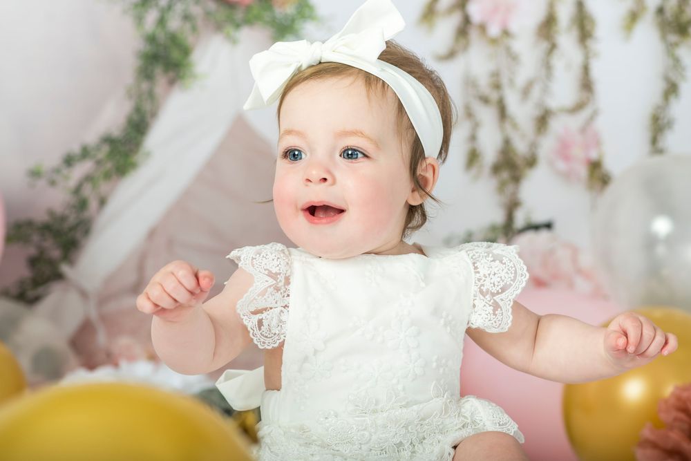 A cheerful baby girl in white lace dress with bow headband smiles at birthday photoshoot.