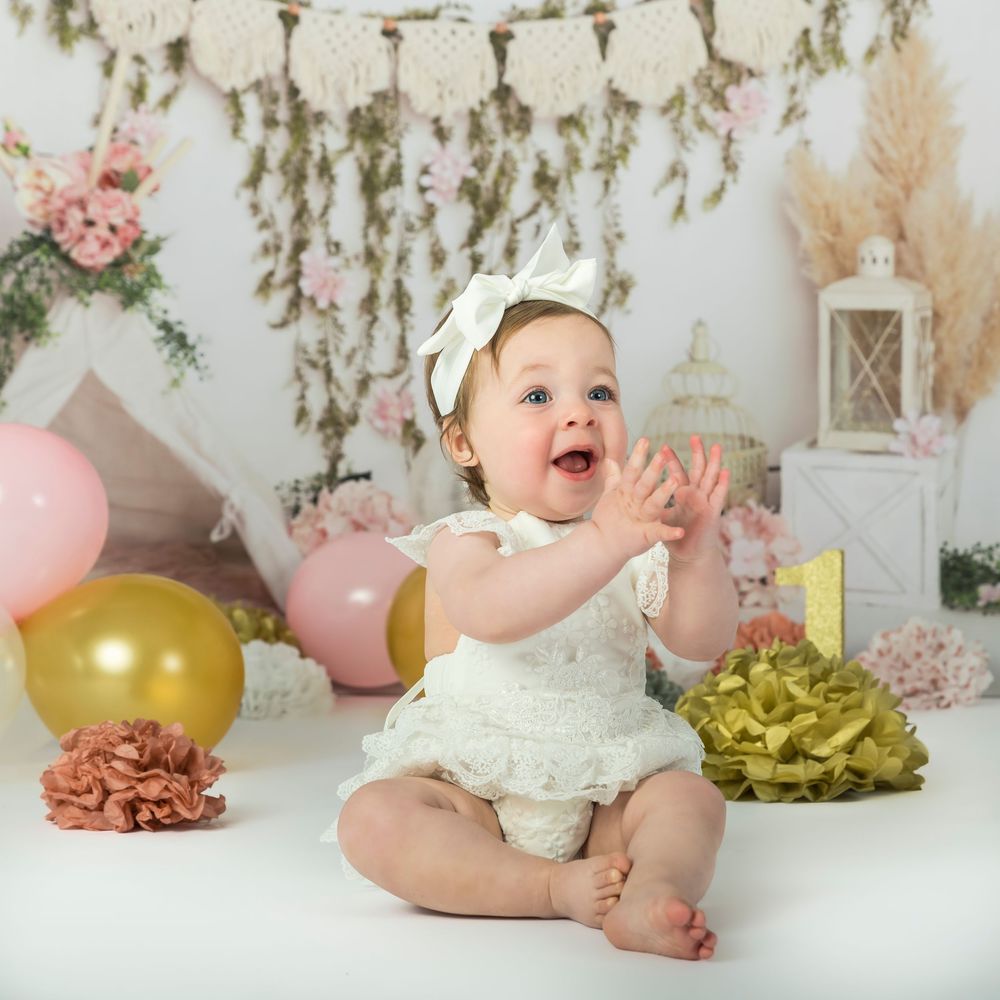 A laughing baby girl in white lace romper claps hands surrounded by balloons and flowers.