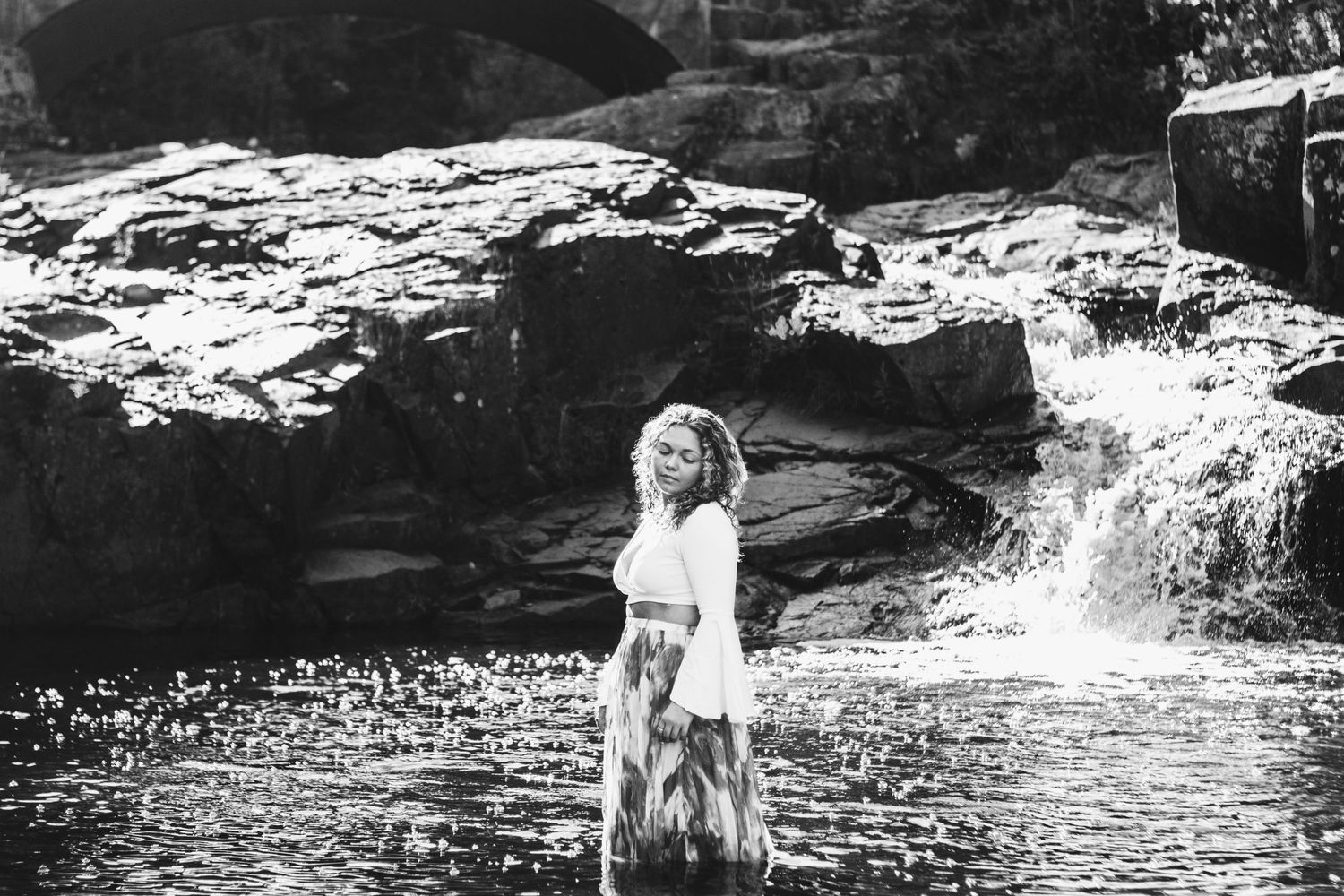 A person in a white dress poses in a shallow stream with a small waterfall and rocky outcroppings in the background.