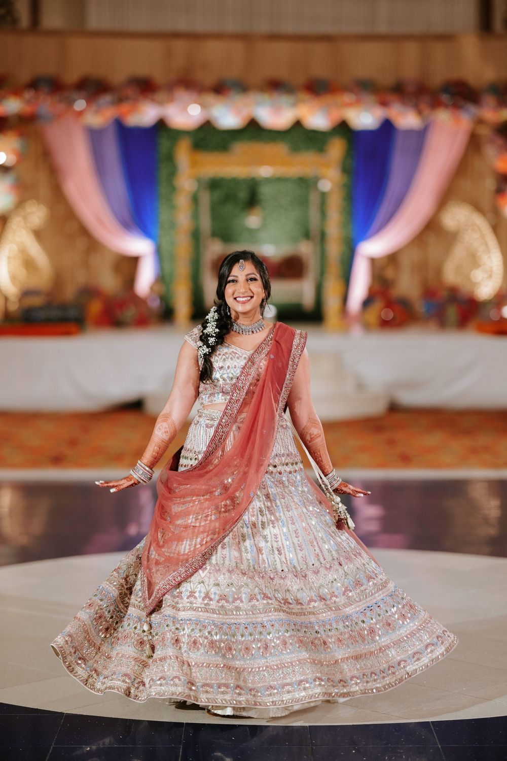 Traditional Indian bridal lehenga in pink and silver with intricate embroidery on a decorated stage with colorful draping.