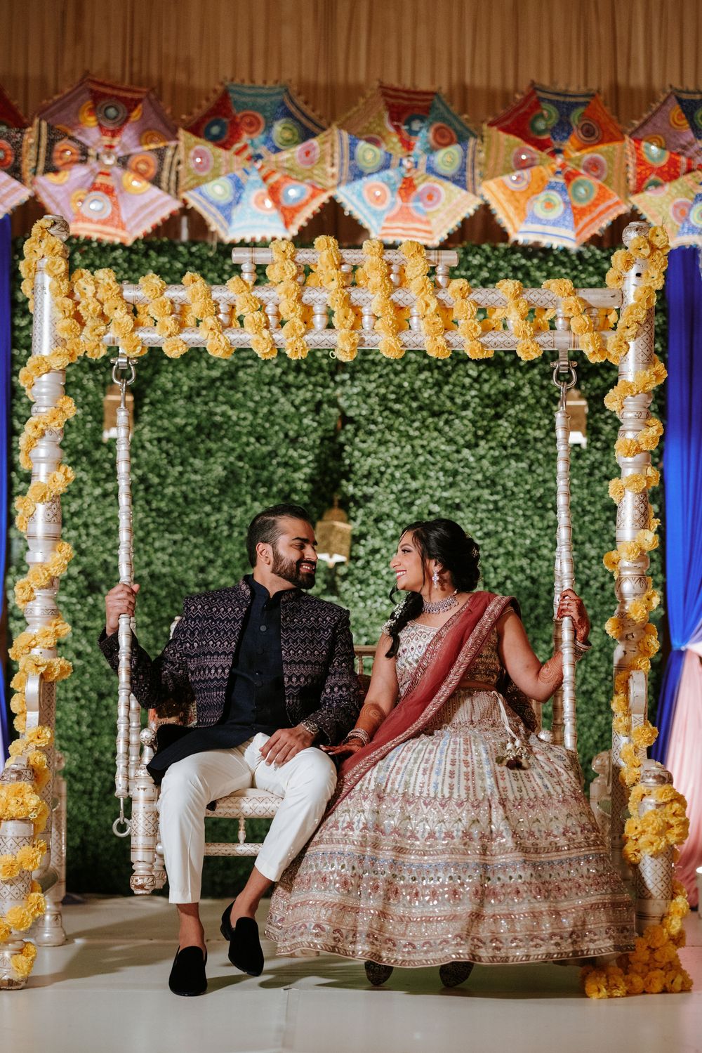 Couple poses together on a decorated swing adorned with floral arrangements and lights at an elegant wedding venue.
