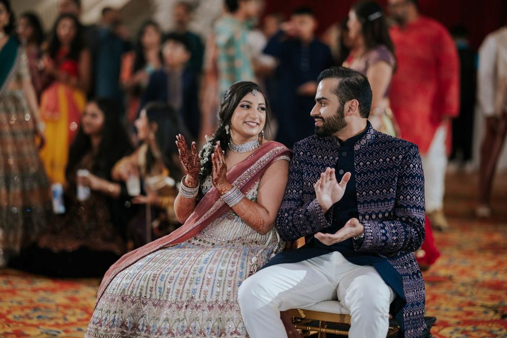 A couple in traditional Indian attire share a joyful moment while sitting at their wedding celebration.