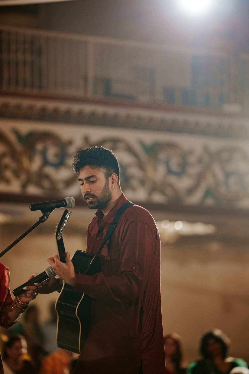 Musician performing with acoustic guitar and microphone at indoor venue in maroon attire.