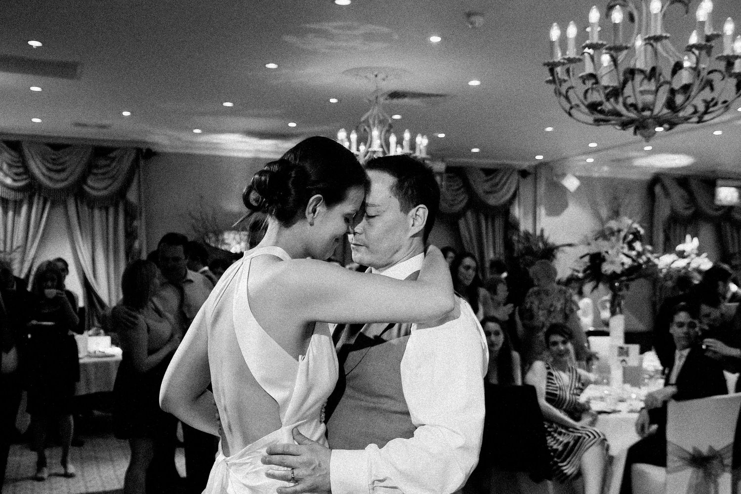 A couple shares their first dance as newlyweds in an elegant ballroom with chandeliers and decorative drapery.