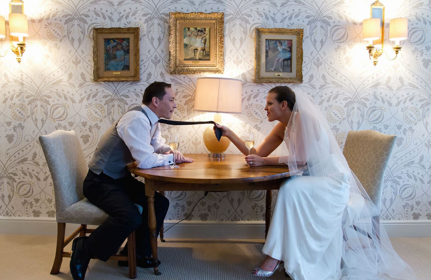 A couple in wedding attire sits at a table arm wrestling in an elegant room with ornate wallpaper and framed artwork.