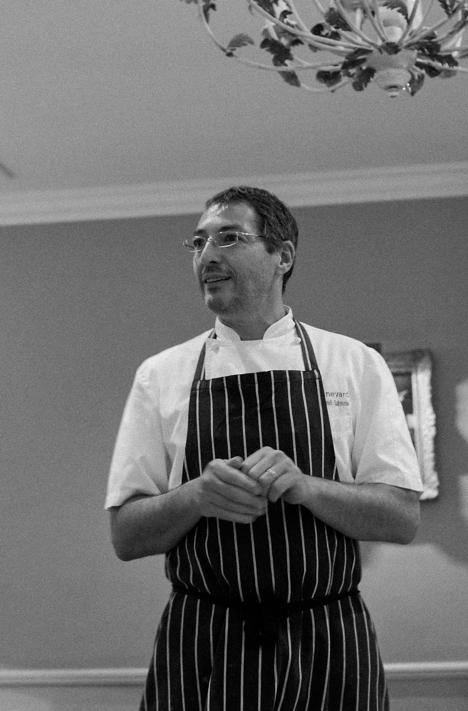 A chef in a white coat and striped apron stands in a kitchen with a chandelier visible above.