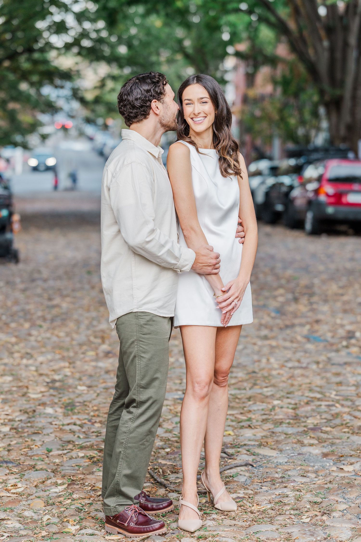 A romantic couple embraces on a cobblestone street during autumn with fallen leaves and parked cars in the background.