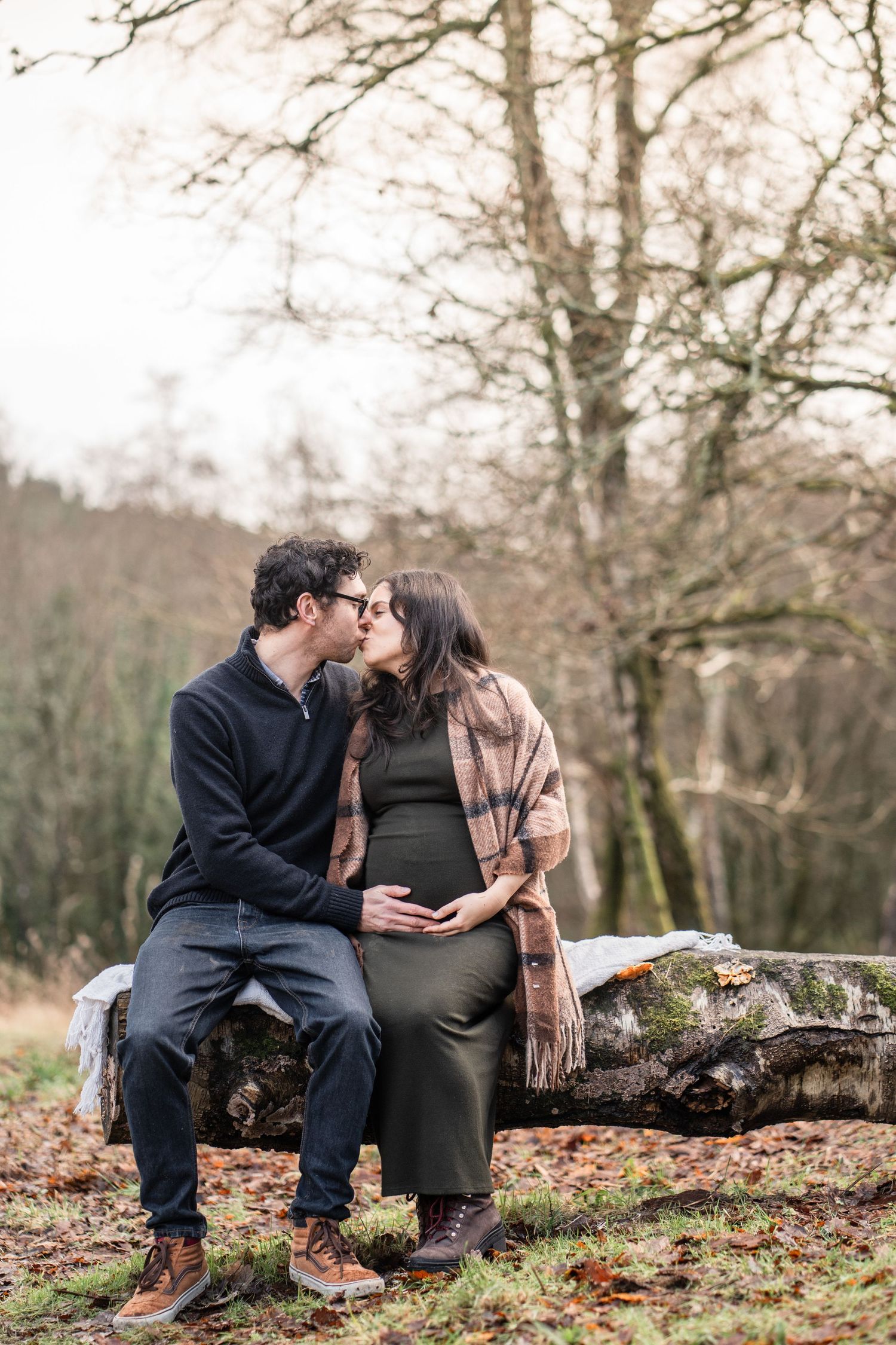 A couple shares an intimate moment while sitting on a fallen tree trunk in an autumn forest setting.