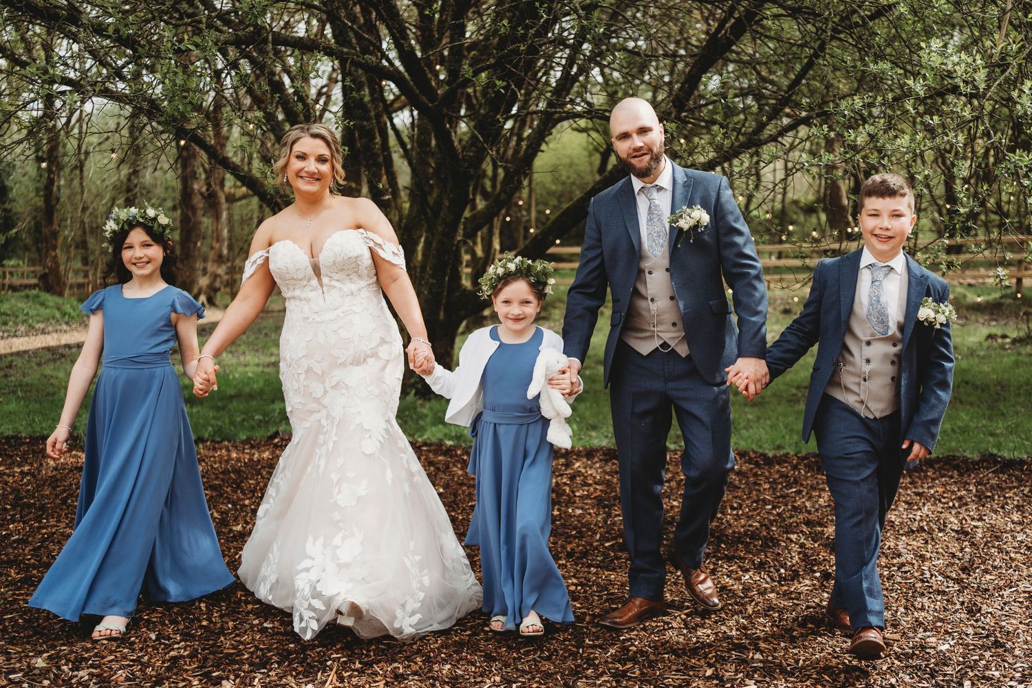 Bride and groom walking hand in hand with their children through woodland, relaxed family moment at a South Wales wedding.